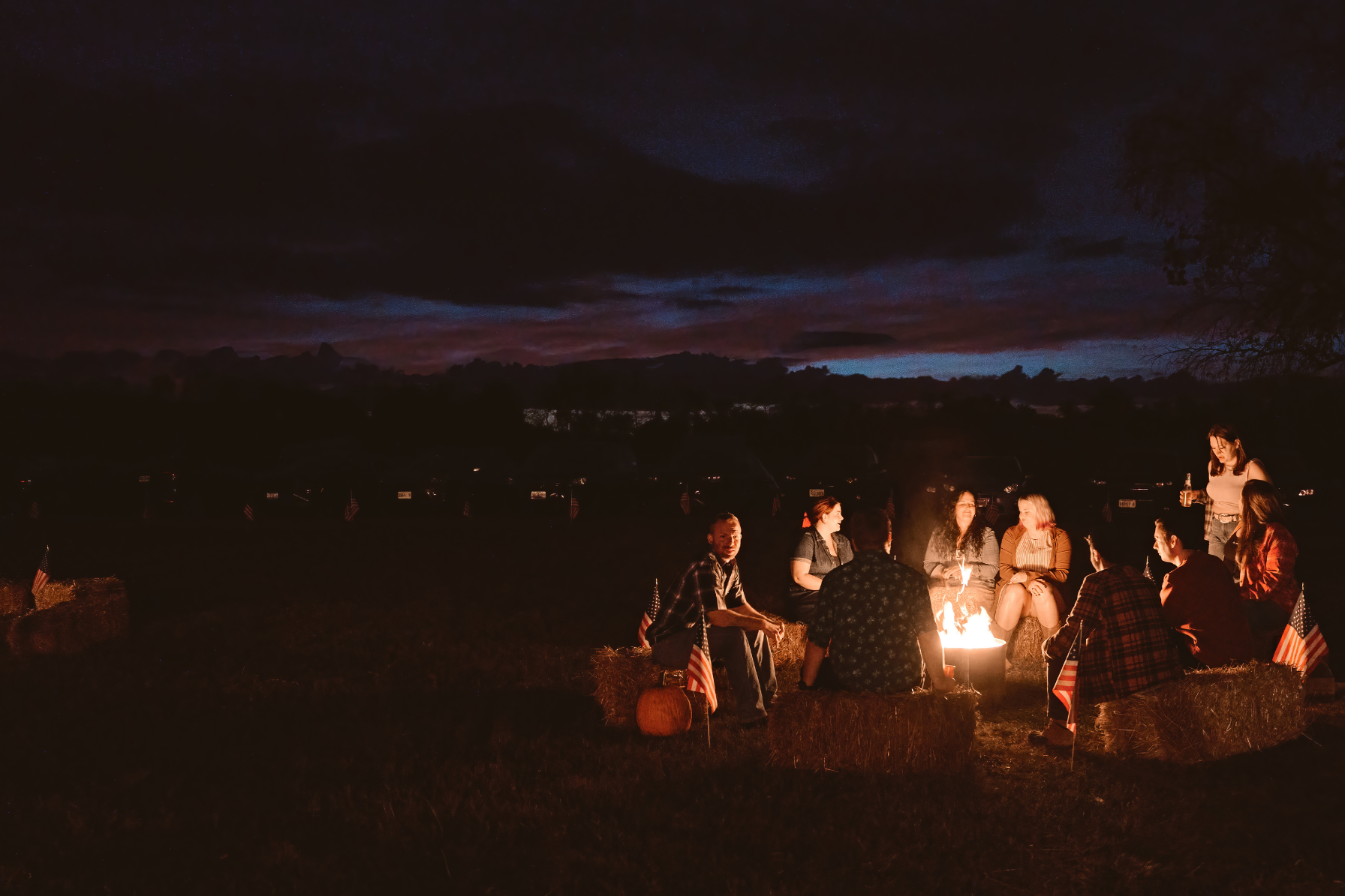 Crowd mingling around a campfire with hay bales, an American flag, and the barn at night